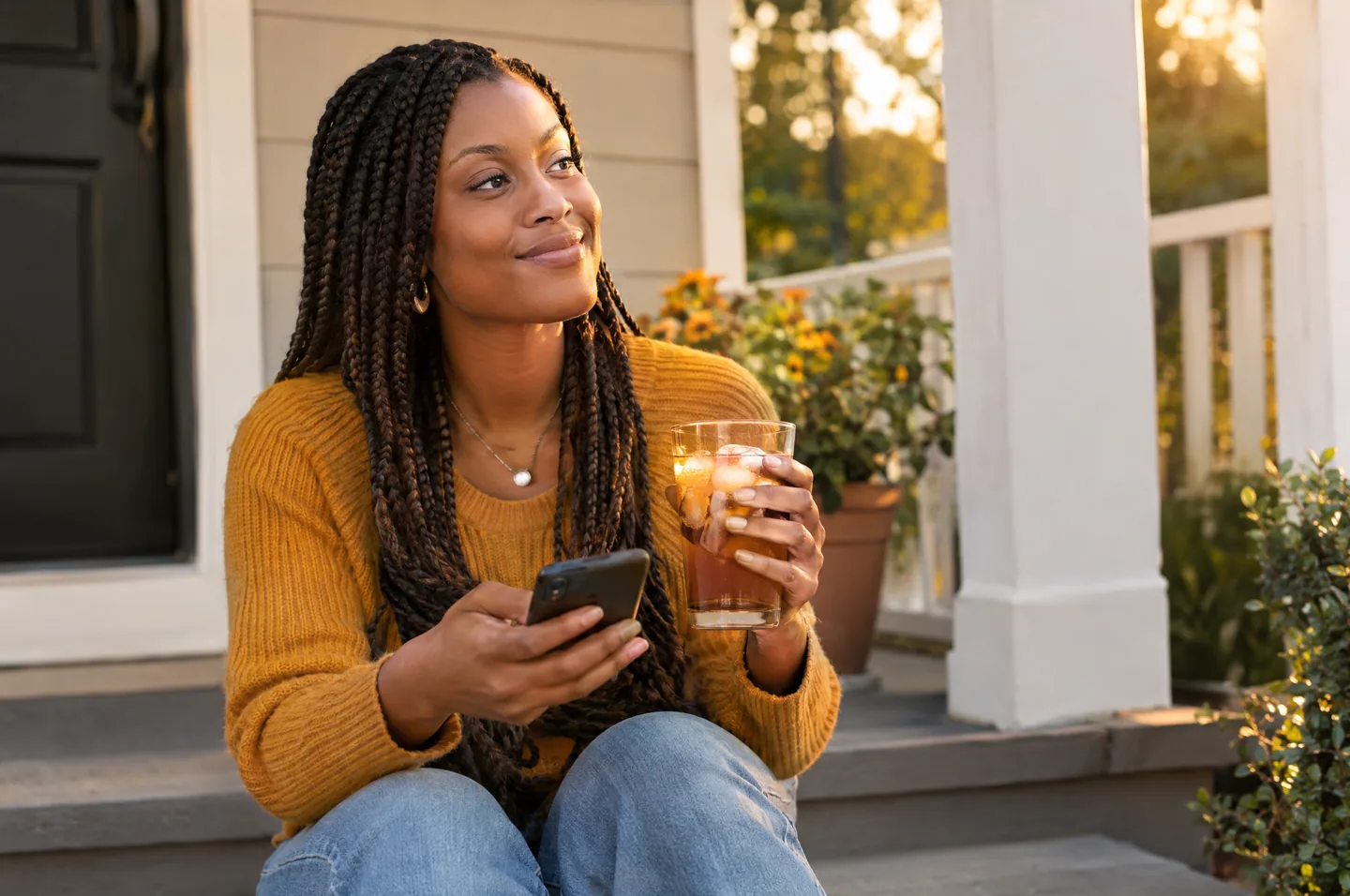 A woman with braids in a mustard sweater sitting on her front porch steps with her phone and a glass of iced tea.