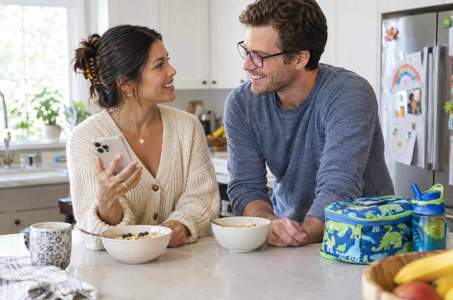 A smiling couple at a kitchen island with cereal bowls and a lunchbox, she holds her phone.