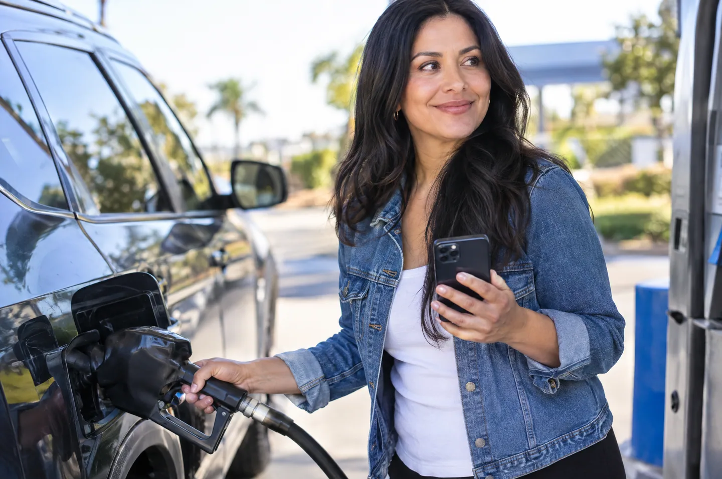 A woman in a denim jacket filling up her car at a gas pump, phone in her other hand.