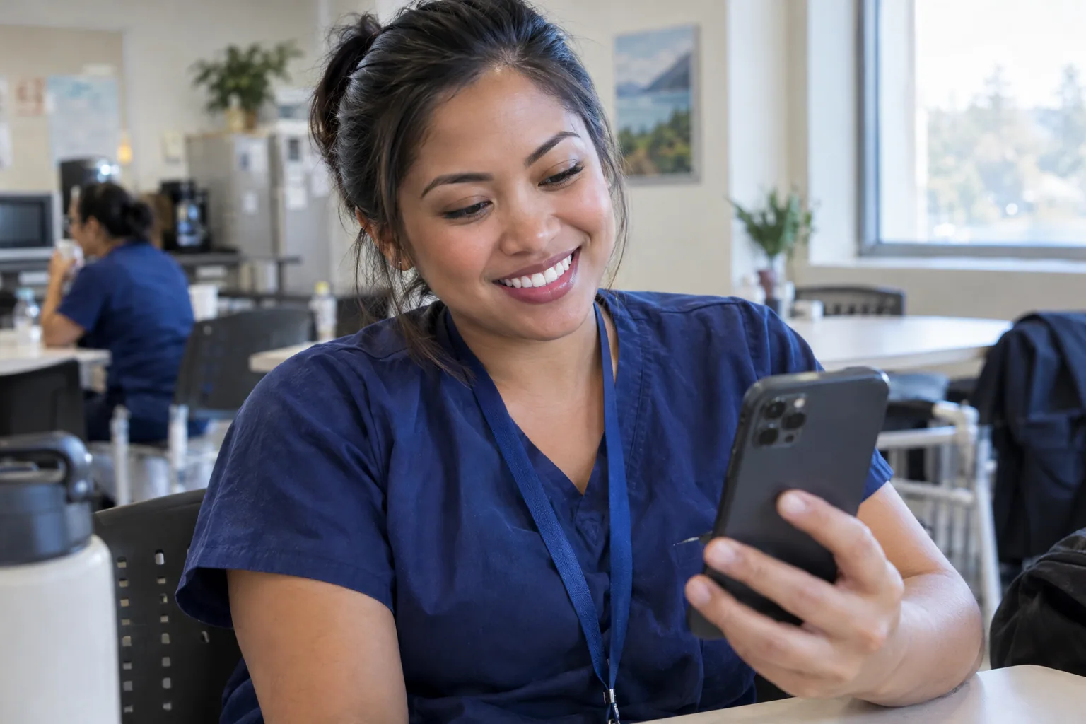 A nurse in navy scrubs in a staff break room, smiling at her phone at the end of a shift.