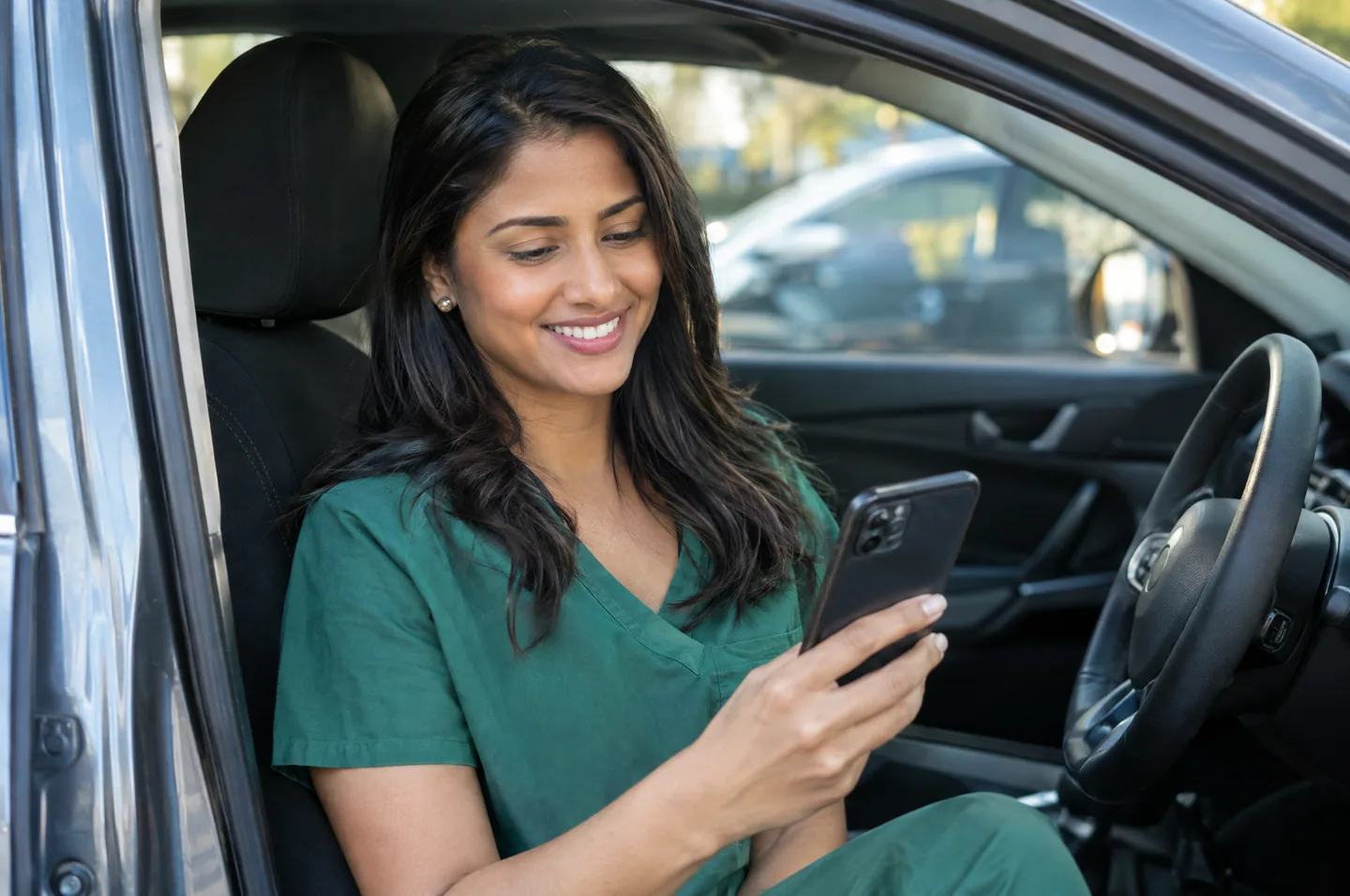 A woman in a green scrub top sitting in her car in a hospital parking lot, smiling at her phone.