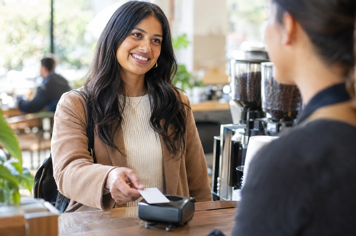 A woman at a coffee shop counter tapping a plain card on a payment reader and smiling at the barista.