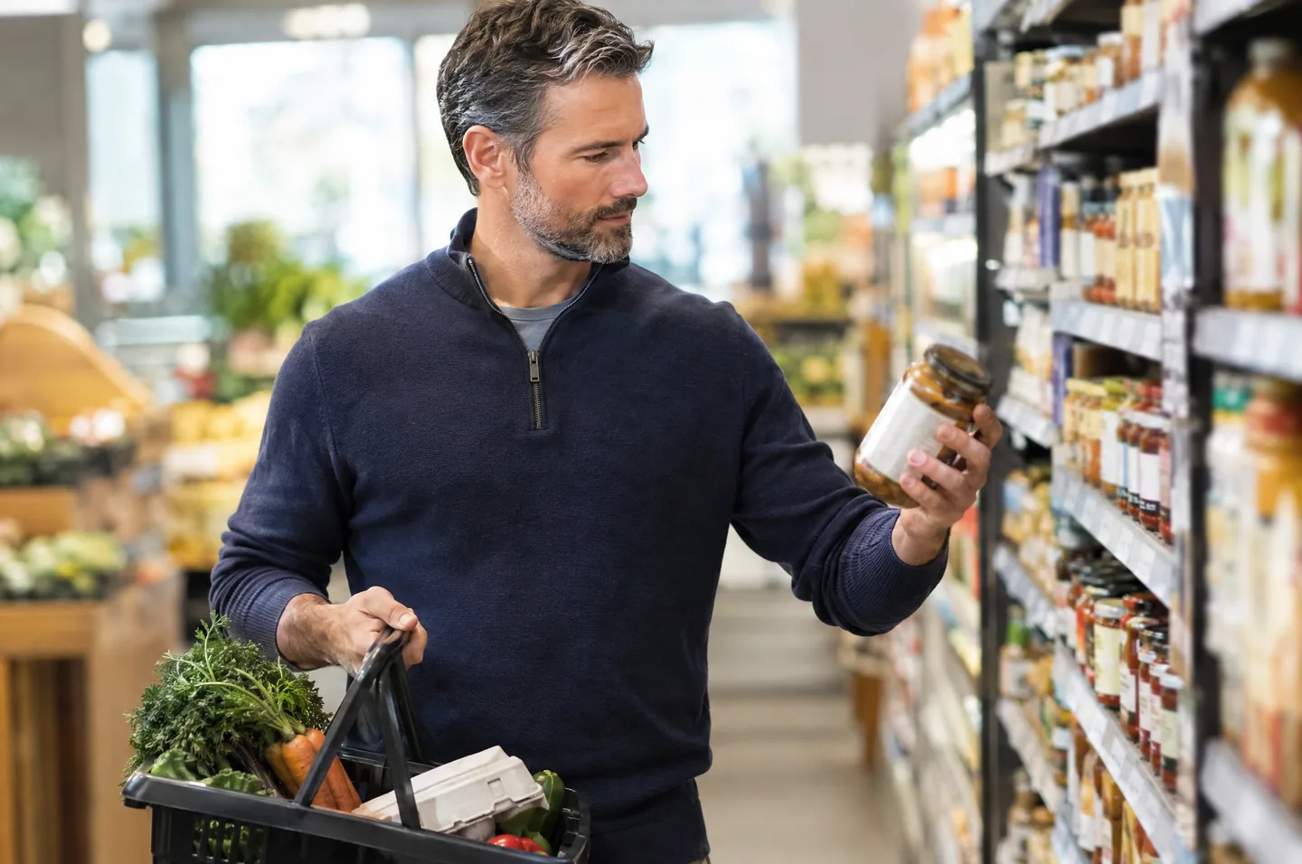 A bearded man in a navy sweater choosing a jar from a grocery shelf, a basket of vegetables on his arm.