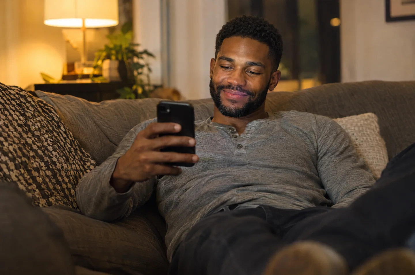 A man in a gray henley relaxing on a living room couch in the evening, holding his phone.