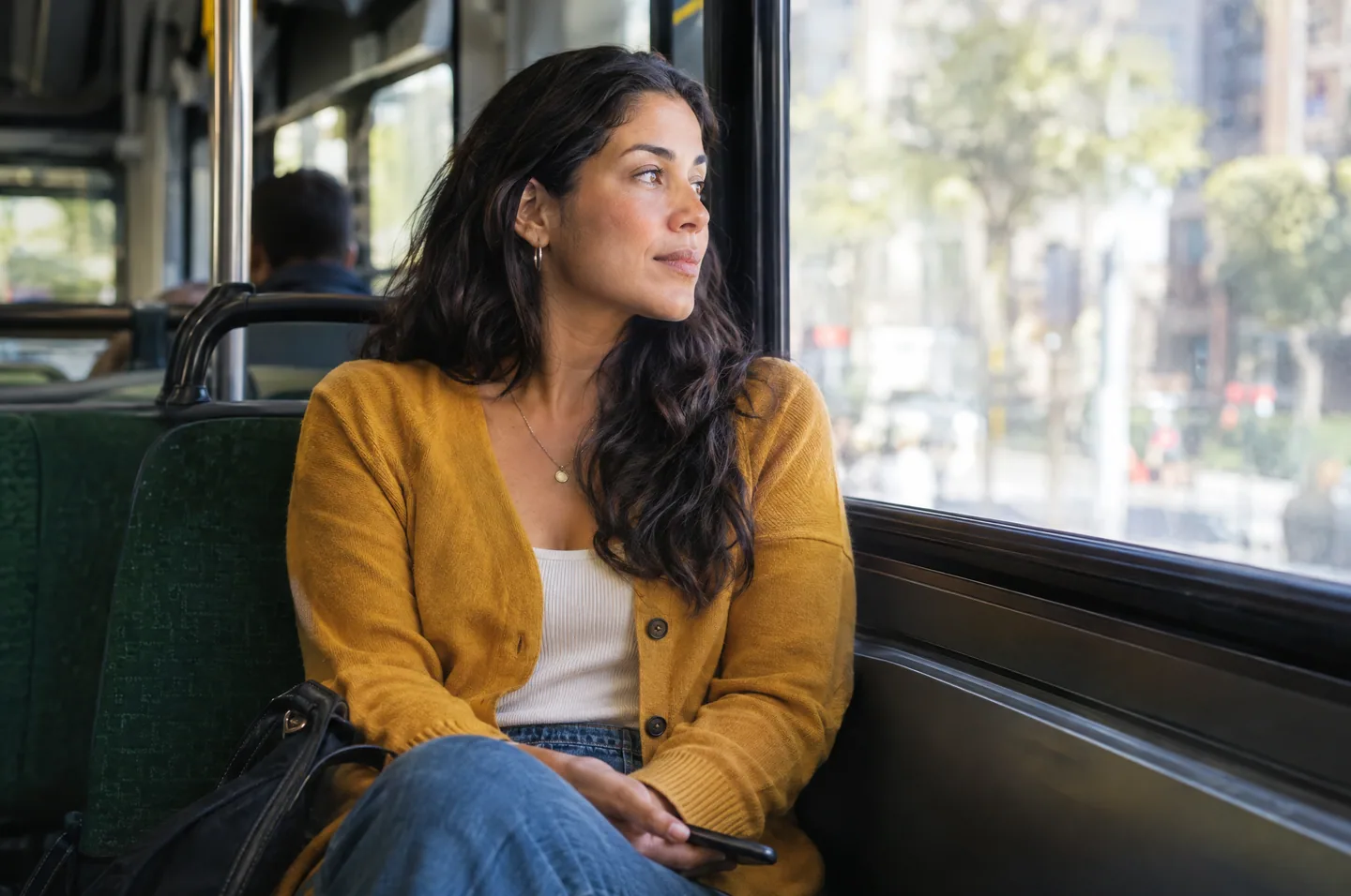 A woman in a mustard cardigan on a city bus, looking out of the window with her phone in her lap.