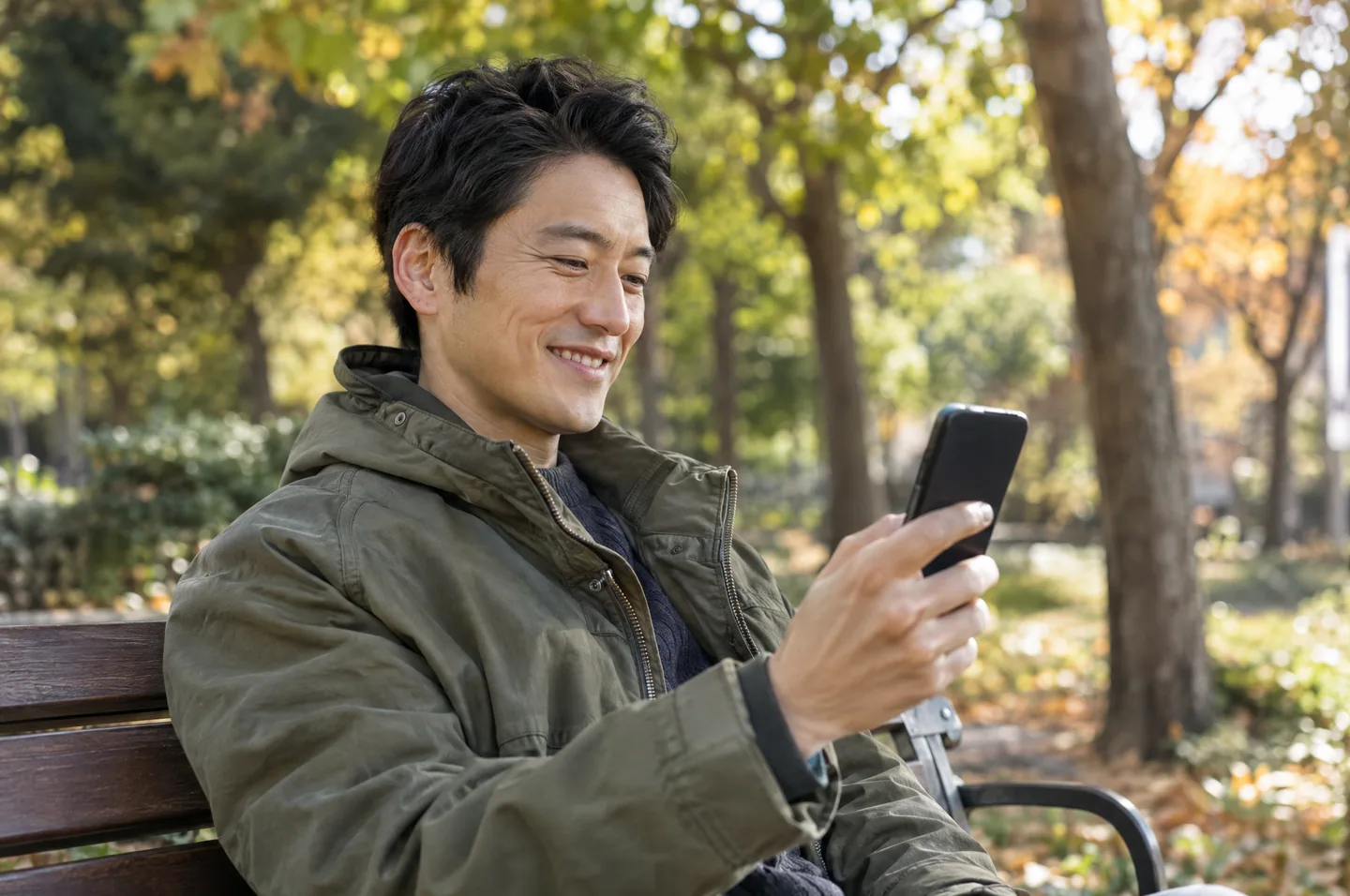 A man in an olive jacket sitting on a park bench on a sunny afternoon, smiling at his phone.